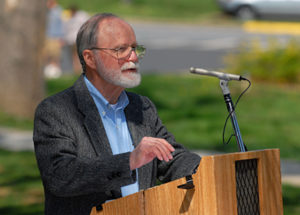 An older white man with grey hair and a white beard. He is wearing a charcoal suit jacket or blazer over a light blue shirt, and he is standing at a wooden dais with a microphone.
