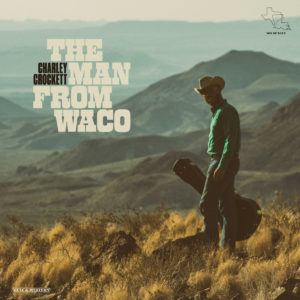 Image: This album cover shows a scrubby mountainous landscape with a Black man in western clothes walking down a slope in the foreground. He wears a cowboy hat, blue shirt, and jeans, and he carries a black guitar case. 
