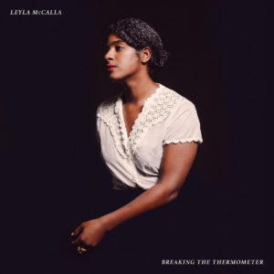 Image: Album cover has a stark black background with a Black woman sitting in the center of it. She has her hair pulled back and faces to the left; she is wearing a white cross-over short-sleeved top and dark pants/skirt.