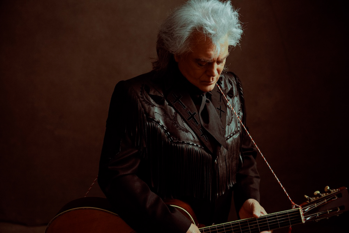A man with grey hair stands against a dark background. He is wearing all black and holds an acoustic guitar.