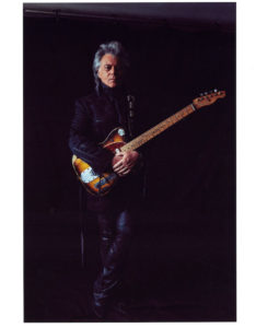 A white man with grey hair stands against a black background. He is wearing all black and holds an electric guitar.