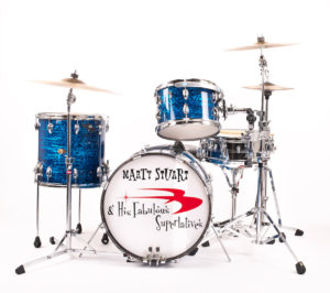 A full drum set against a white background. The drums are a deep blue color and the large drum in the front has the band name -- Marty Stuart & His Fabulous Superlatives -- on its head. Several cymbals can also be seen.