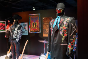 One of the exhibit displays from The World of Marty Stuart. Several stage costumes can be seen, along with posters and prints in the background. There are also a several instruments on display.