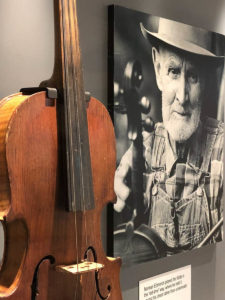 Close up of a fiddle hanging in a display case. A black-and-white photograph of an older bearded white man is seen to the side of the fiddle. He is holding a fiddle and he wears a checked shirt, overalls, and a hat.