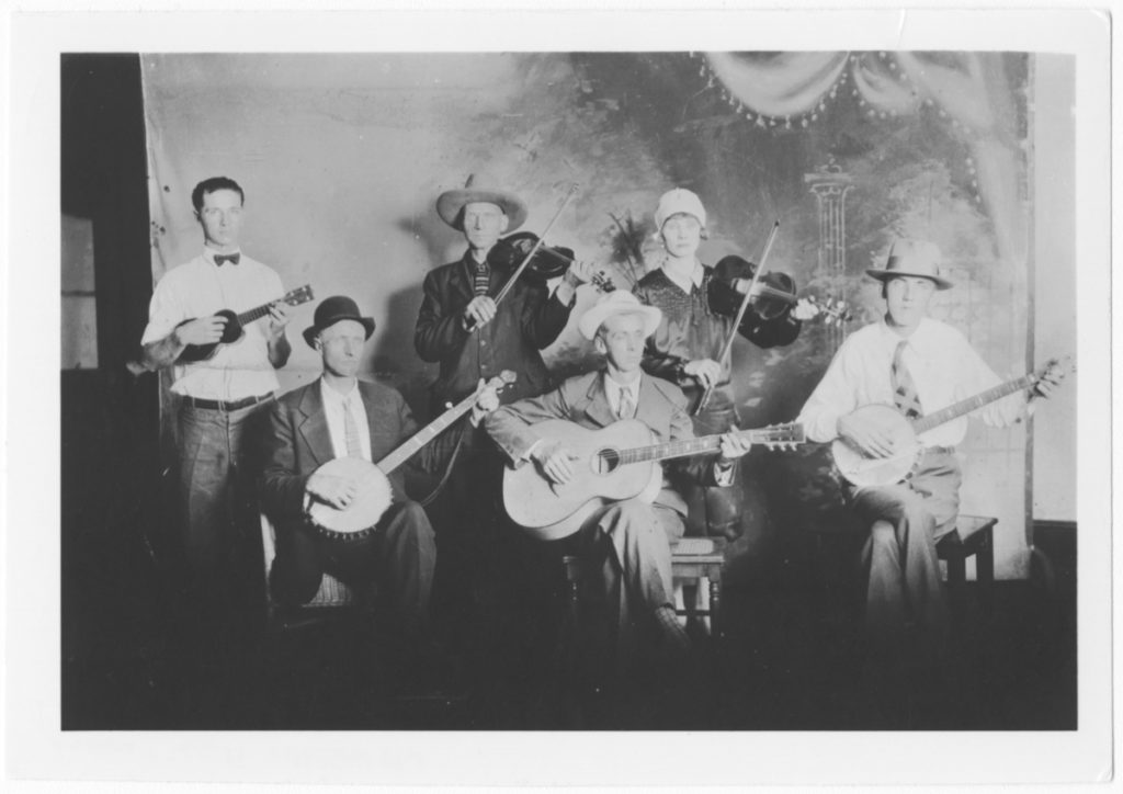 String band posed in front of a false backdrop. Three white musicians stand at the back: a ukulele player and two fiddle players, one of which is a woman. Three musicians sit at the front: two banjo players and a guitar player.