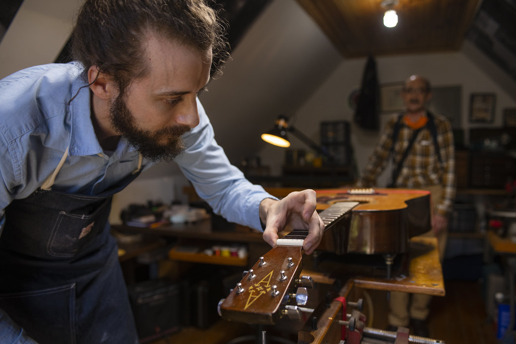 A white man with dark hair and beard leans over the neck of a guitar. He is working on the inlay design. In the background, an older white man stands and watches his work.