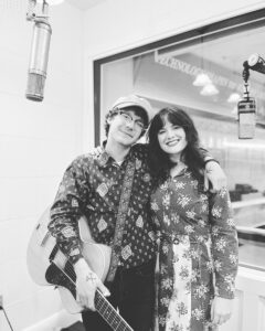 Black-and-white photograph of two musicians. The male musician is to the left -- he is white, wearing a hat, glasses, and a patterned button-down shirt. He holds a guitar. The female musician is to the right, with long dark hair and wearing a patterned dress. They are in a radio studio and you can see two microphones to either side and the glass window of the booth behind them.