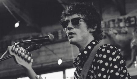 Black-and-white photograph of white male musician on stage -- he is playing a guitar and singing into a mic. He has dark curly-ish hair, and wears sunglasses and a spotted/patterned shirt.