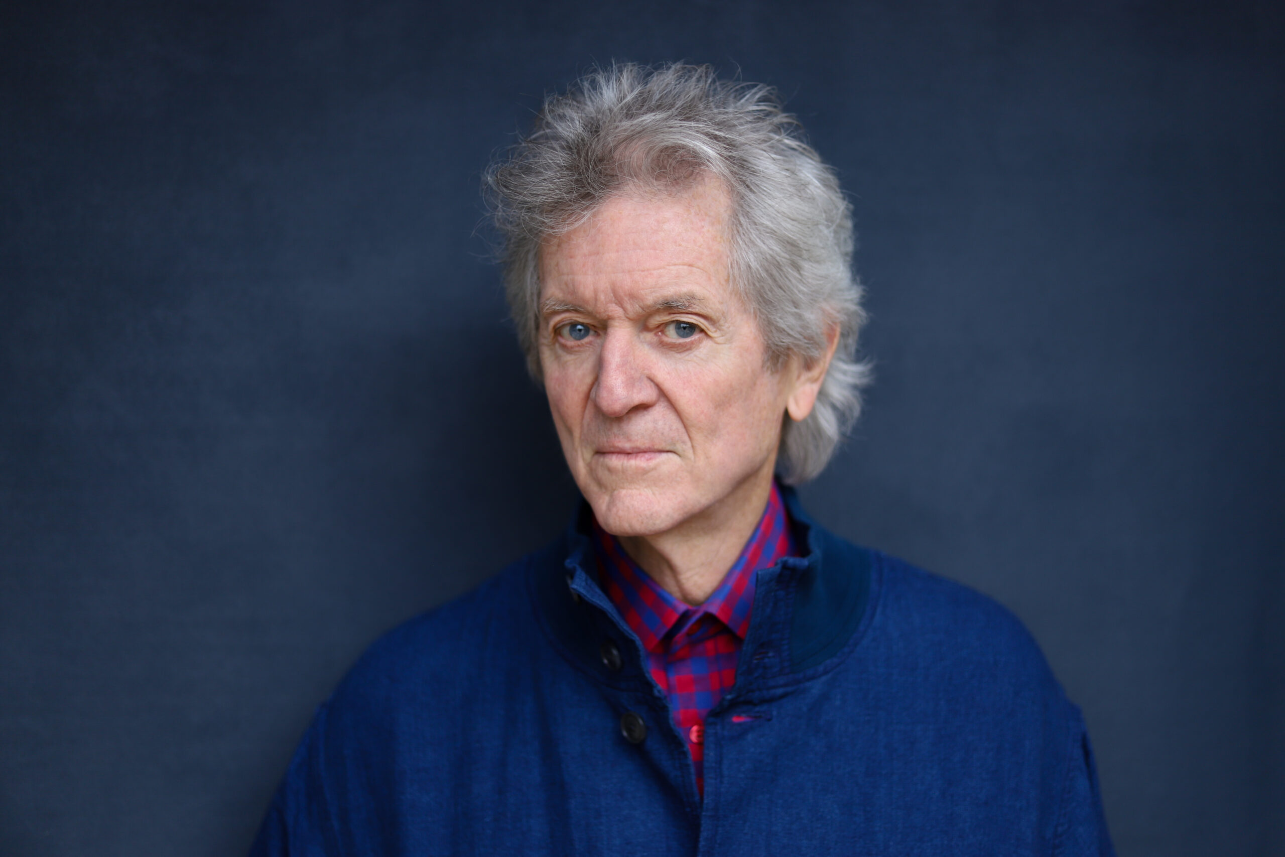 A head shot of Rodney Crowell standing in front of a wall.