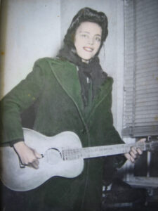 A photo of Etta Baker in her youth, wearing a coat and head scarf and playing her guitar.