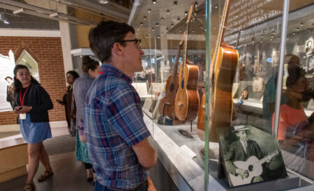 A group of teachers visit the museum during an in-service workshop. In the foreground, a white male teacher with dark hair, glasses, an da blue plaid shirt looks at a guitar in the display case.