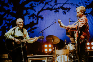 97 year old Clyde Lloyd looks onward toward Tyler Childers as they play onstage. Both are playing guitars in front of a stage backlit by a blue backdrop with a large silhouette of a tree behind them. 