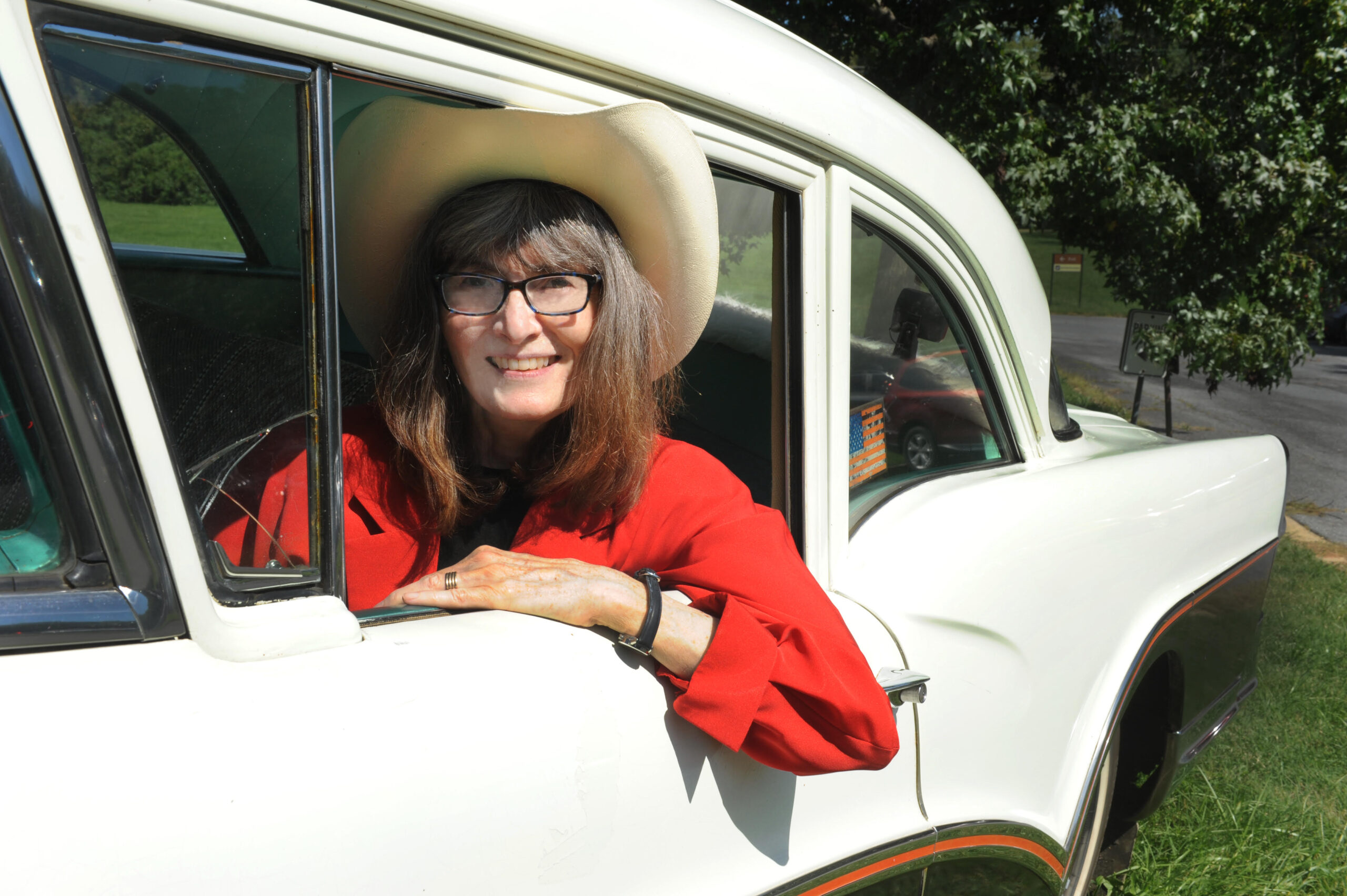 Karen Collins, wearing a cowboy hat, and sitting behind the wheel of a classic car.