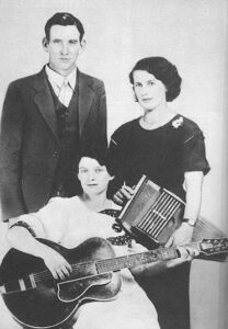 A black and white image of The Carter Family. Three people are facing the camera, A.P. Carter is wearing a blazer and vest, looking toward the camera. Sarah Carter is to his left and is standing facing the camera. She is holding an autoharp and wearing a dress. Maybelle Carter is sitting holding an archtop guitar and looking into the lens. All three individuals have a slight smile to their faces. 