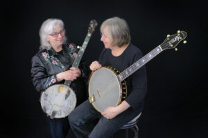 Two white women sit in front of a black background. Cathy (to the left) has white shoulder length hair and is wearing a black and embroidered western-style shirt; she holds a banjo. Marcy (to the right) has chin length grey hair and is wearing black; she also holds a large banjo.