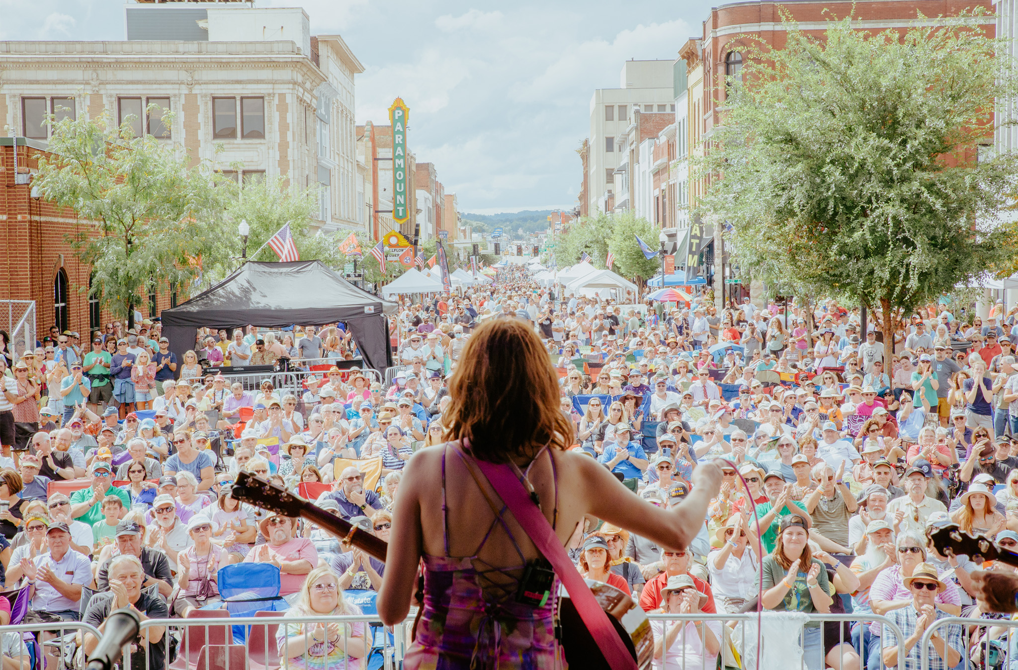 A photo taken during Bristol Rhythm & Roots Reunion music festival depicting a woman standing on stage, holding a guitar. Her back is to the camera as she is facing a sea of several thousand people as she performs for them on stage.