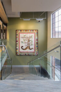 A large colorful quilt is shown hanging on a display on a wall inside of the Birthplace of Country Music Museum. The image shows the quilt on display over a staircase during the daytime with dim spotlights highlighting the quilt. 
