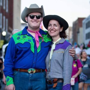 A man and a woman standing in a street and looking at the camera. The man is white and wearing jeans, a royal blue and green western-style shirt with a red bandana around his neck, sunglasses, and a cowboy hat. He has his arm around a white woman. She is wearing a checked and purple western-style shirt, jeans, and a brown hat.