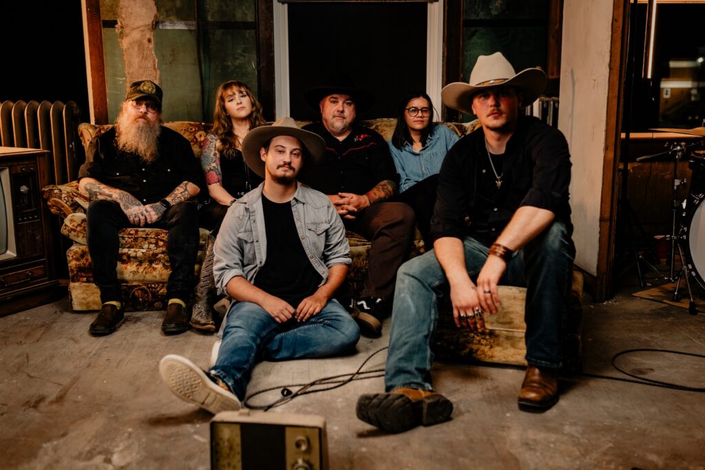  A promotional image of the group The Dimestore Cowboys. A group of six musicians are posing all facing the camera and looking into the lens. They are sitting on an old 1970’s style floral couch next to an old large TV with amps and other miscellaneous music equipment surrounding them. The lighting is moody and dark, no one is smiling. Members of the group are dressed in rustic western attire and dark clothing. 