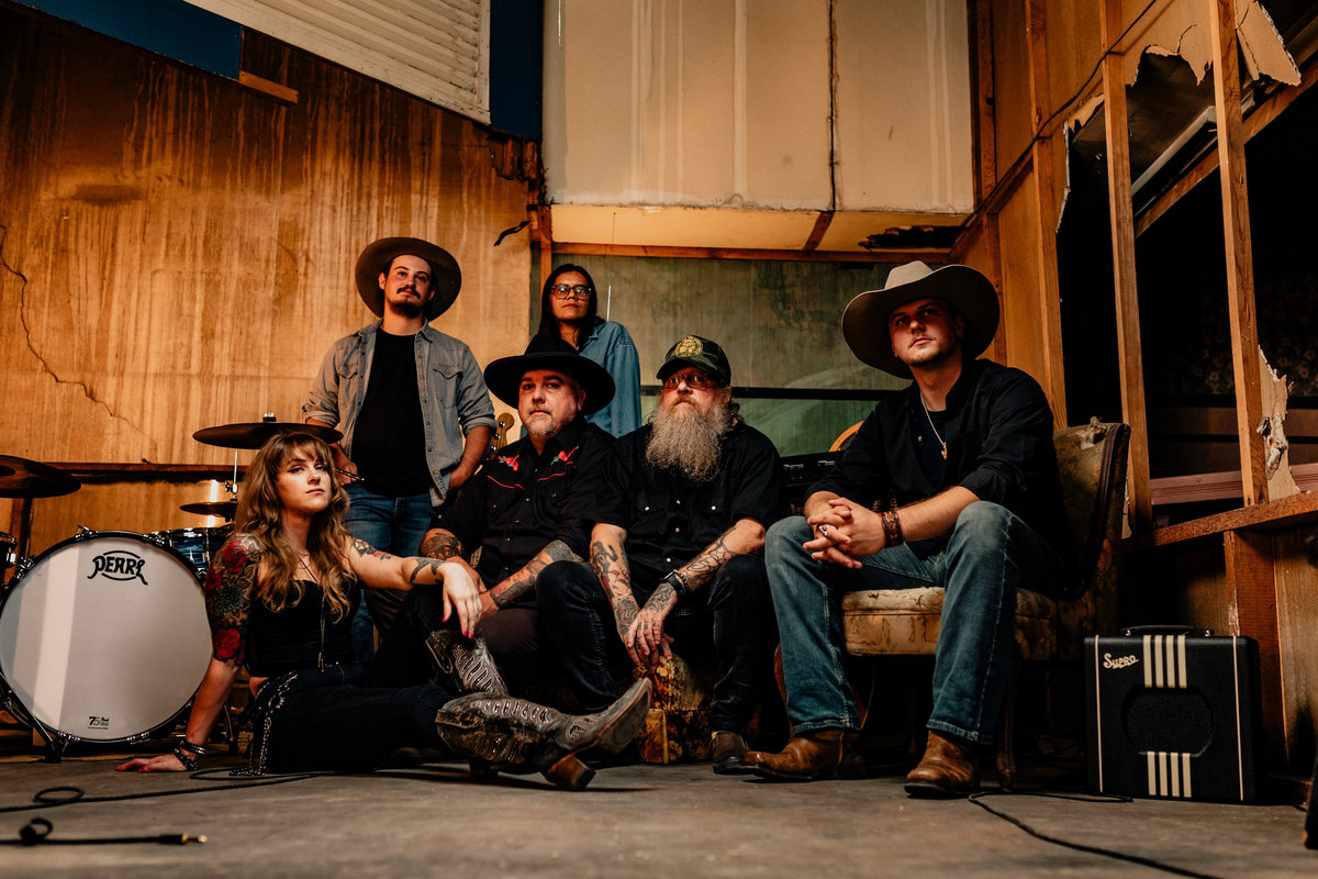 A promotional image of the group The Dimestore Cowboys. A group of six musicians are posing all facing the camera and looking into the lens. They are all posed in front of drum kit and amps in a studio space. The lighting is moody and dark, no one is smiling. Members of the group are dressed in rustic western attire and dark clothing.