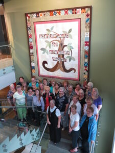 A large group of twenty four women standing on a staircase inside of the Birthplace of Country Music Museum in front of the handmade quilt. The camera is facing to the right of the group of women and many of them are smiling. 