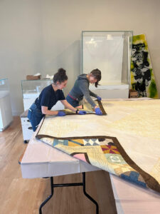 Two museum workers are leaning over multiple folding tables covered in white plastic and are carefully folding the birthplace of country music museum quilt. Julia Underkoffler is to the right of Rene Rodgers; both are smiling and wearing blue rubber gloves and handling the corners of the quilt. Julia is wearing a navy blue shirt and blue jeans and Rene is wearing a gray hooded sweatshirt and blue jeans. They are standing in a gallery room with museum display cases behind them.