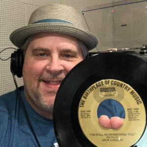 A selfie of Big Lon inside of the Radio Bristol studio space holding a record with that reads "The Birthplace of Country Music Bristol" on the front. Big Lon (Lonnie) is smiling and wears a fedora straw hat and studio headphones. 