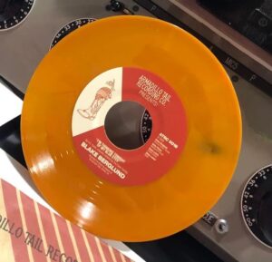 A closeup of a bright orange colored vinyl 45rpm record. The text on the front of the record reads "armadillo tail recording company presents" and the bottom text reads "Blake Berglund" in larger lettering with smaller text. An armadillo with his head inside of a cowboy boot is also on the record. 