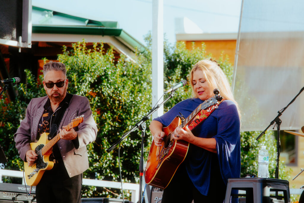 A photo of Carlene Carter singing and playing an acoustic guitar alongside Chris Casello on electric guitar. Photo is taken at Bristol Rhythm & Roots Reunion 2023 Country Music Mural stage.