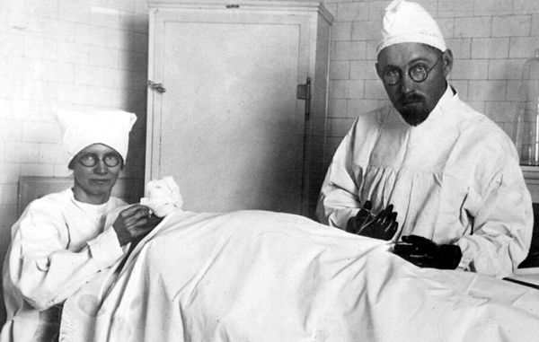  a black and white image of a women and a man looking directly at the camera performing surgery on a person laying on a table with a white sheet placed over their body. The women is wearing a surgical hat, and round glasses. The man is to her right and also wearing a surgical hat and round glasses, and is holding a surgical tool in his hand and gloves