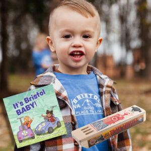 A photo of a toddler aged boy wearing a Bristol sign t-shirt with insets at the bottom of photos of a wooden train whistle and a little book entitled "It's Bristol Baby."