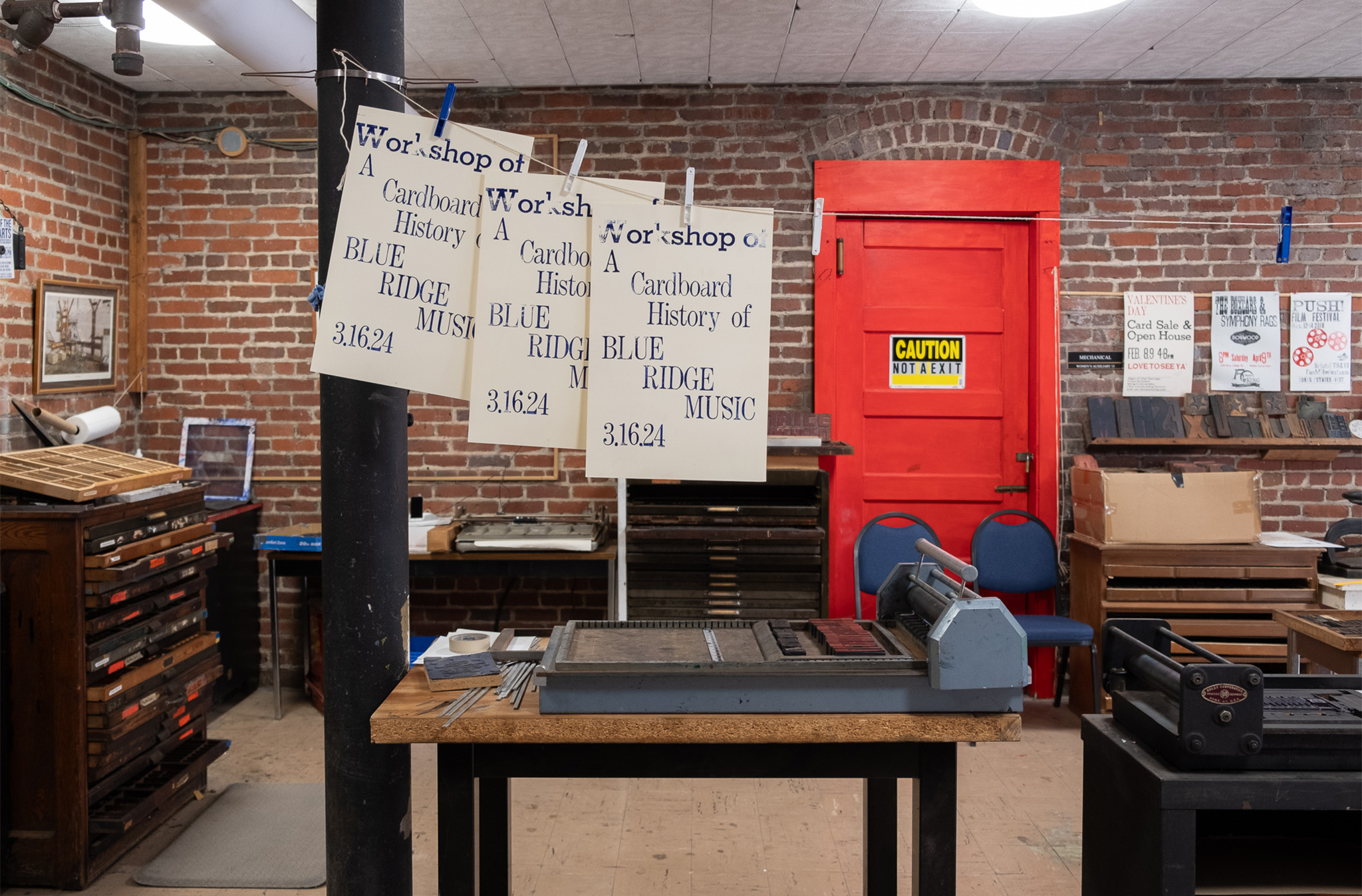A brick-walled room with a table and letterpress printing equipment. There is a red door on the room's back wall and several letterpress printed folders hanging on a clothesline above the table. The posters including information about the March 16 letterpress workshop.