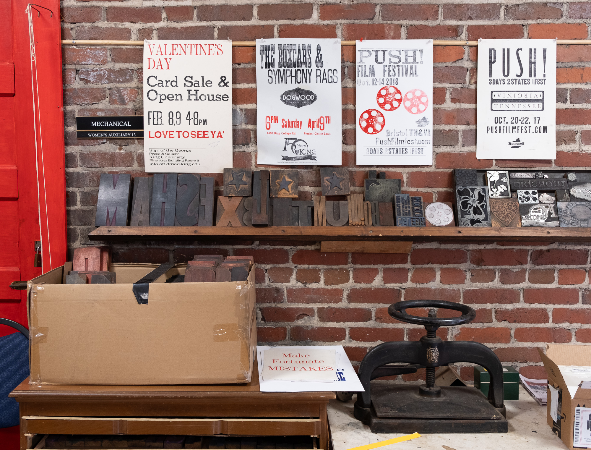 A scene inside of a letterpress print shop. Posters are hung in a line against a brick wall. Wooden letters and type face are on a shelf under the posters. A black hand press and box full of type is on the table under the shelf.