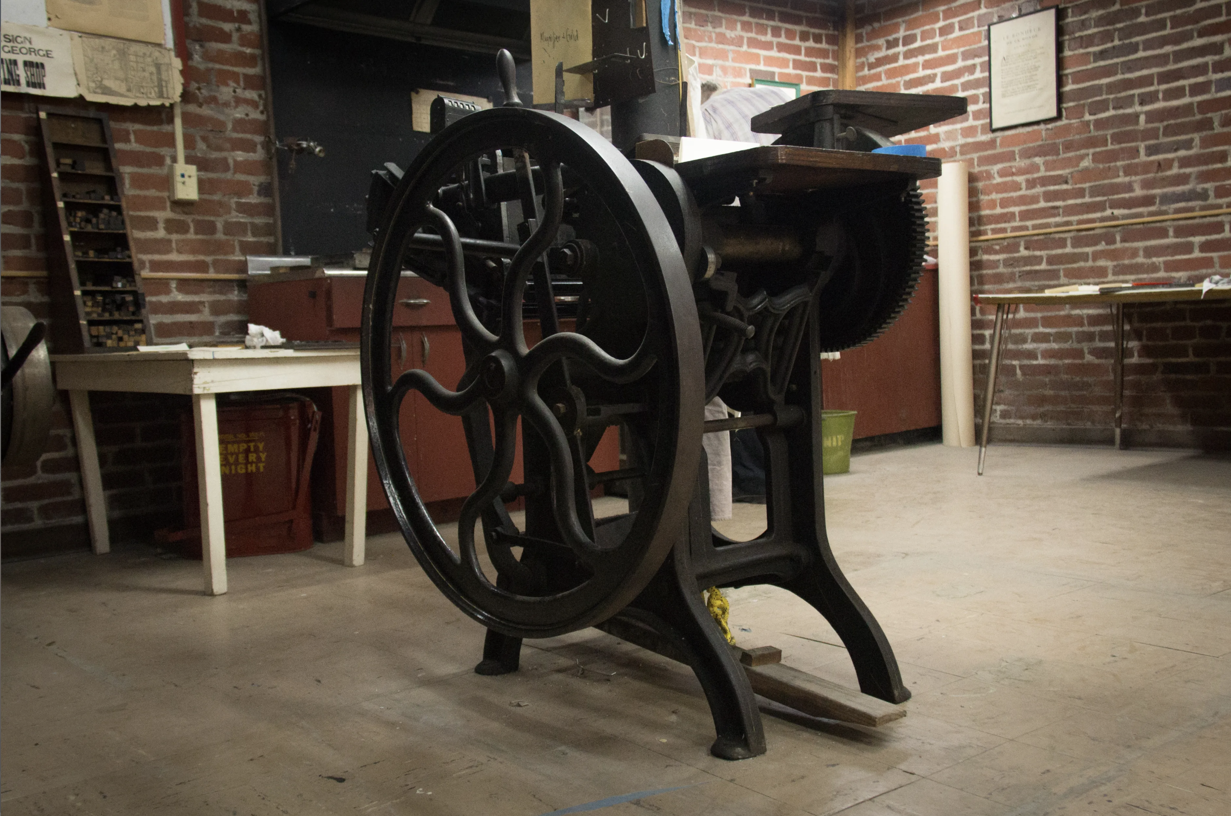A large black printing press inside of the print shop, a brick wall is surrounding the space. 