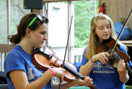 A photo of two teenaged girls playing fiddles; they are wearing JAM Kids t-shirts.