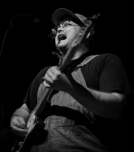 A white man in a baseball cap and overalls playing a guitar while singing.