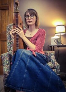 Woman with short brown hair and glasses wearing a pink cardigan and jean skirt sits in a floral chair with crossed legs and holding a dulcimer upright in her lap.