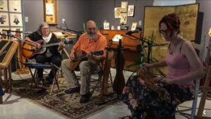 Three people play instruments together in chairs in front of mics. The woman on the left is playing a guitar. The man in the middle is playing a mandolin. The woman on the right is playing a dulcimer.