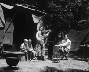 A white man holding a baby plays a piano outside a trailer while a standing woman plays fiddle and two children sit on a bench beside her. 