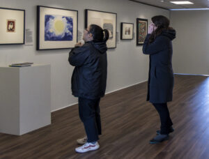 Two people in dark winter clothes stand in a narrow gallery looking at various artworks on the wall.