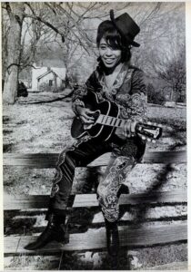 A black woman in a rhinestone studded outfit sits on a fence playing a guitar and smiling.