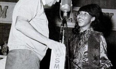 A Black woman smiling and standing next to a mi with WSM Grand Ol Opry written on it while a white man adjusts the mic for her.