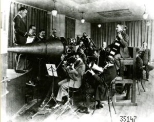 An image of men playing in an acoustic recording session. Multiple musicians sit crowded around a large metal horn, playing directly into it so it captures their sound.