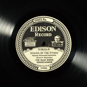 An old record labeled "Edison Record: The Sinking of the Titanic, Singing, Harmonica, and guitar. Ernest V. Stoneman, The Blue Ridge Mountaineer." On the sides of the label are an image of a laboratory with the words "A product of the Edison Laboratory" under it, and an image of Thomas Edison.