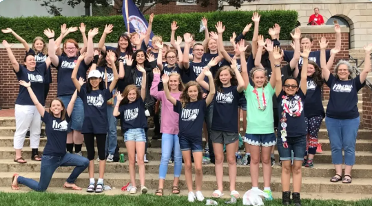 A large group of students, mostly white, stand on a set of stairs. The majority of them are wearing dark blue Virginia History Day t-shirts. The kids all have their arms in the air with excitement.