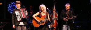 An image of Emmylou Harris, standing between two other musicians with her guitar. She has white hair, is smiling, and lightly strumming the guitar.