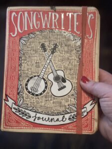 A photo of a red book titled "Songwriter's Journal." It had a drawing of a crossed guitar and banjo on the cover.