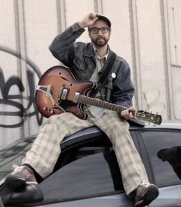 A photo of Matty Sheets sitting on the roof of a car, holding his guitar in his lap and tipping his ballcap with his hand.