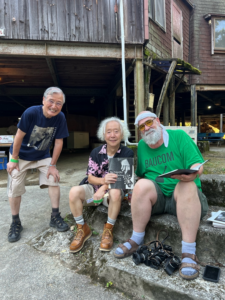 Three elderly men, two Japanese and one Caucasian, posing together for a photograph. The man in the center is holding up a book of bluegrass photography.