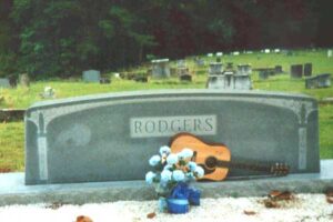 Large headstone with the word "Rodgers" in the middle. with a pot of white flowers and a guitar laid in front.